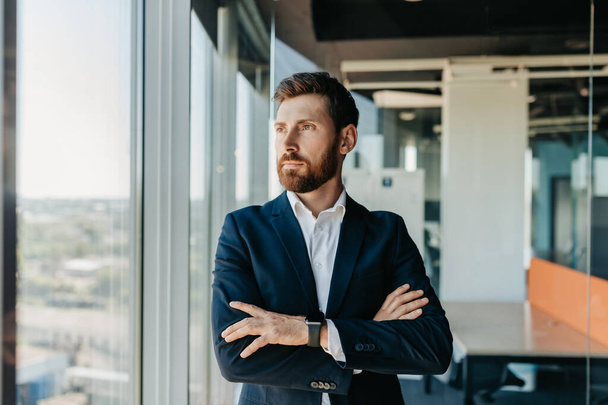 Successful businessman in formal wear standing with folded arms in modern coworking office and looking through window, thinking about startup, planning new projects, waiting for meeting - Foto, afbeelding