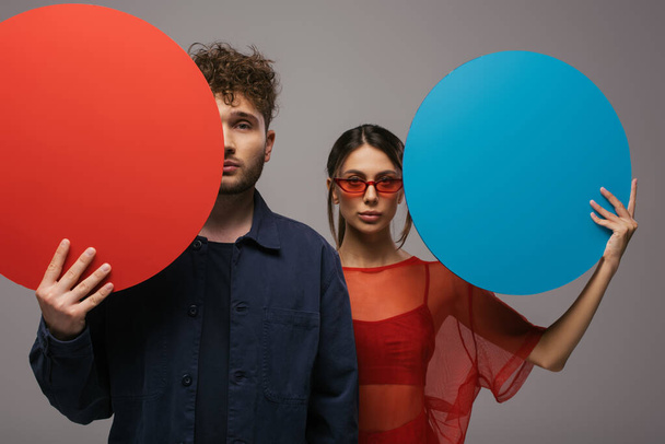 young couple in blue and red outfits holding round shape carton and looking at camera isolated on grey - Foto, imagen