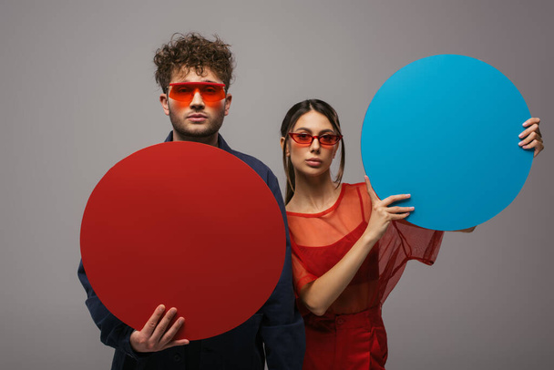 young couple in blue and red outfits and sunglasses holding round shape carton isolated on grey - Foto, Imagen