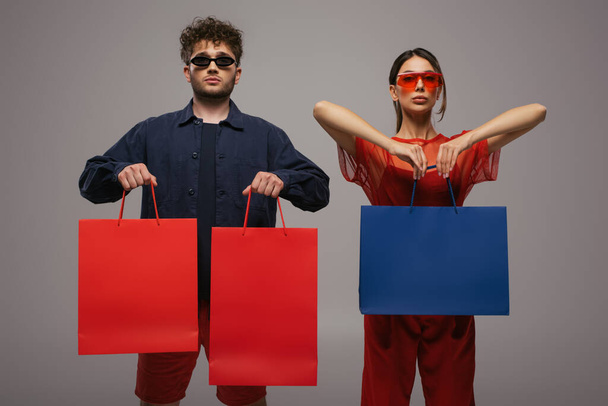 young couple in trendy outfits and sunglasses holding shopping bags isolated on grey - Foto, Imagem