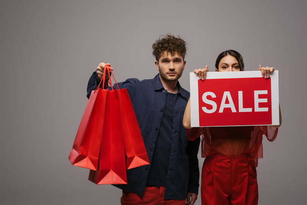 young couple in trendy outfits holding shopping bags and paper with sale lettering isolated on grey - Φωτογραφία, εικόνα