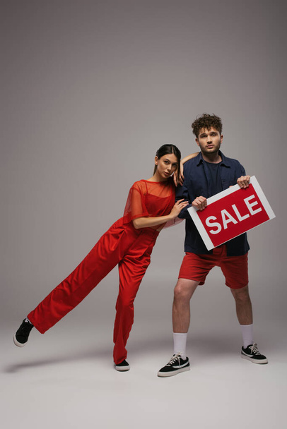 full length of young couple holding paper with sale lettering on grey - Foto, Imagem