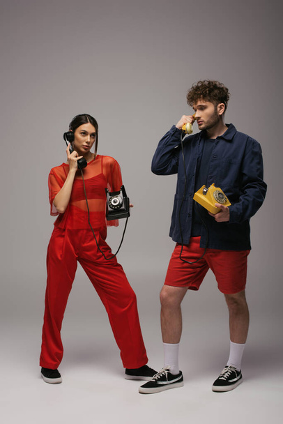 full length of young woman and curly man holding retro handsets and telephones on grey - Photo, Image