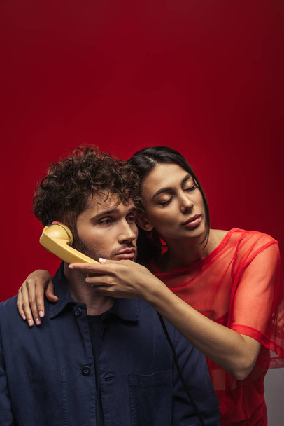 young woman holding retro handset near ear of curly man on red - Foto, Imagen