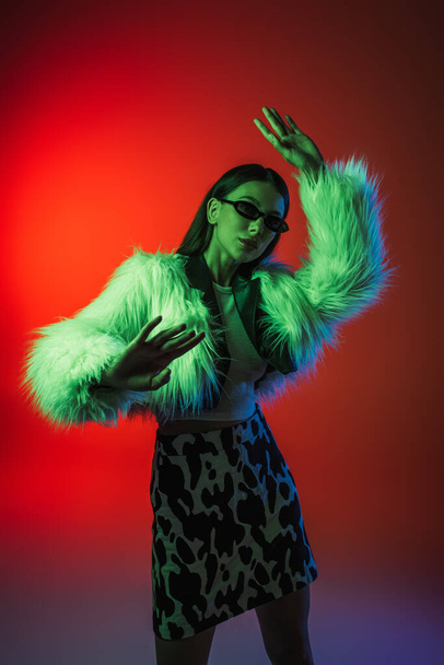 stylish young woman in faux fur jacket and sunglasses gesturing while posing on red - Photo, Image