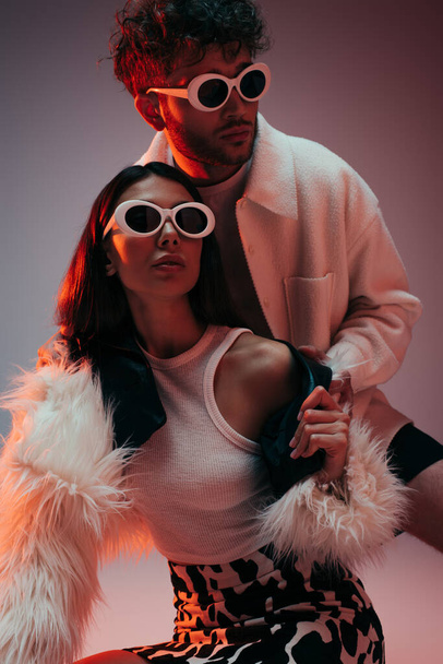 curly man and stylish woman in sunglasses and faux fur jacket posing on grey - Fotó, kép