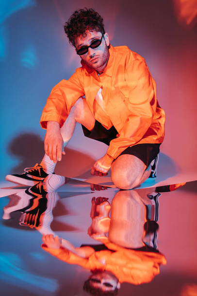 full length of curly young man in sunglasses posing while sitting on blue with red lighting  - Zdjęcie, obraz