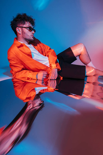 stylish and curly young man in sunglasses posing on blue with red lighting  - Φωτογραφία, εικόνα