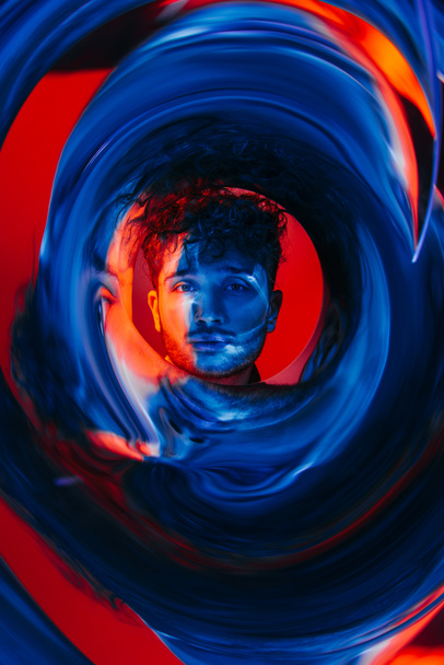 curly man looking at camera through futuristic circle on red and blue background  - 写真・画像