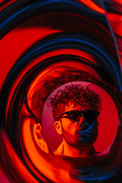 curly man in sunglasses looking away through futuristic neon circle on red - Foto, Imagen