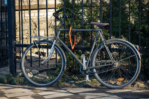 Чорний ретро-велосипед припаркований на фоні металевого паркану з стіною будинку та садом на задньому плані. Літній, екологічний, популярний транспорт. Вінтажний велосипед з замком. Безпечна парковка в місті
 - Фото, зображення