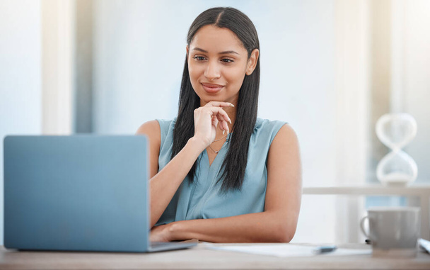 Young mixed race business woman holding hand on chin while working on laptop. One female entrepreneur smiling while reading an email or getting an idea while looking at laptop screen in her office. - 写真・画像