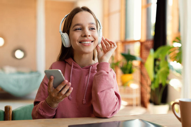 Boldog kaukázusi tinilány vezeték nélküli fejhallgatóval és mobiltelefonnal, otthon ül az asztalnál és mosolyog. Tinédzser hallgat zenét vagy hangoskönyvet, amelynek távoli webes lecke - Fotó, kép