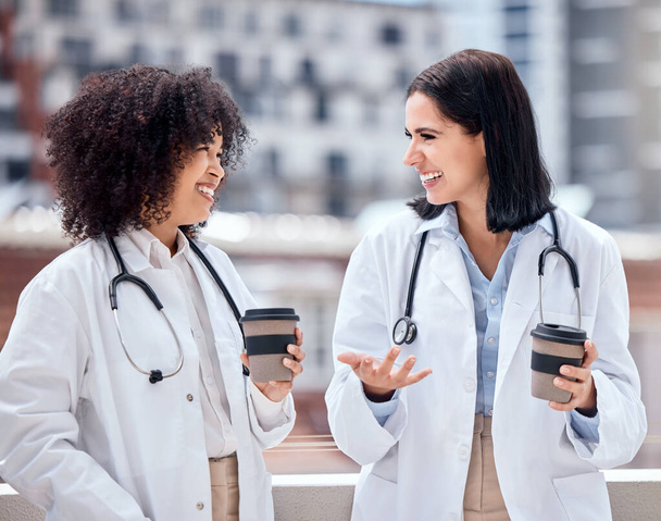 Deux femmes médecins heureuses et gaies debout et buvant une tasse de café dehors faisant une pause ensemble. Femmes hispaniques de race blanche et mixte souriant relaxant et parlant sur une pause à l'extérieur. - Photo, image