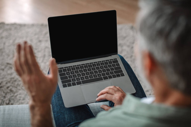 Przycięty starszy biały mężczyzna macha ręką i patrzeć na laptopa z pustym ekranem, ma czat we wnętrzu pokoju, przez ramię widok, zbliżenie. Technologia dla emerytów w domu, rozmowy wideo i spotkania zdalnie - Zdjęcie, obraz