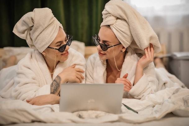 Bornozlu güzel genç kadınlar ve yatakta laptoplu ve kahveli havlular. - Fotoğraf, Görsel