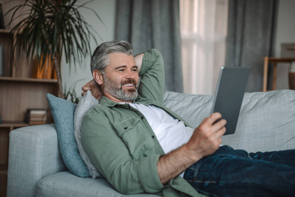 Cheerful old caucasian man resting on sofa and watching video on tablet in living room interior, copy space. Device for pensioner at home, new app for relax, chat in social networks in free time - Foto, immagini