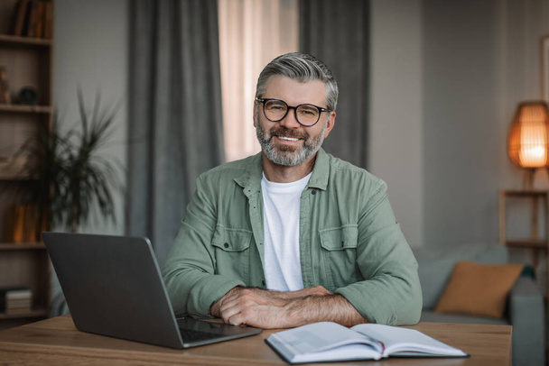 Glad mature caucasian male teacher with beard in glasses working with laptop, shooting video lesson in room interior. Tutoring with modern device and study at home, social distancing and work remote - Foto, Imagem