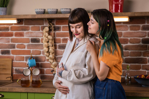 Good looking lesbian couple very enthusiastic standing beside the country style kitchen they waiting a baby young lady partner caresses the belly of her pregnant girlfriend concept of LGBT freedom - Photo, image
