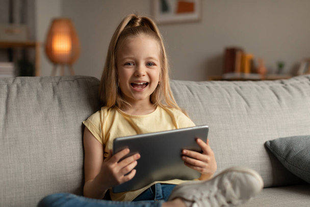 Menina alegre rindo usando tablet digital sorrindo para câmera sentada no sofá em casa. Criança se divertindo navegando na Internet e jogando jogo on-line no computador. Crianças e Gadgets - Foto, Imagem