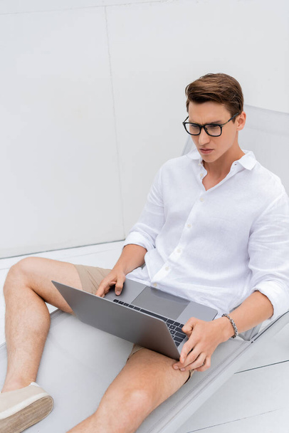 thoughtful man in white shirt and eyeglasses using laptop in deck chair outdoors - Foto, Imagen
