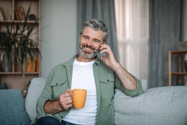 Glad mature european man with beard calls by smartphone, enjoys favorite hot drink in living room interior. Social distance and communication, device for retired at home, rest and relax at free time - Foto, Imagem