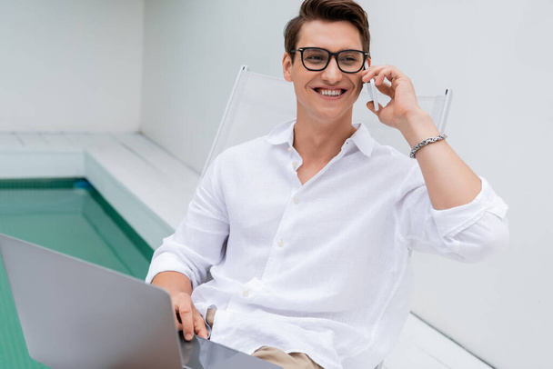 cheerful man talking on smartphone near laptop and pool while looking away - Photo, Image