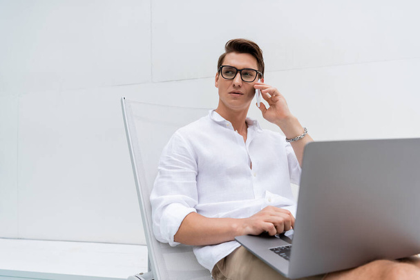 freelancer em óculos sentado em cadeira de praia e falando no celular perto do laptop - Foto, Imagem