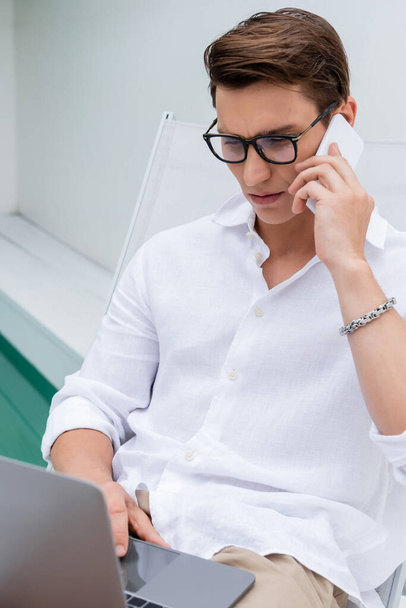 freelance concentrato in camicia bianca e occhiali da vista utilizzando il computer portatile e parlando sul cellulare all'aperto - Foto, immagini