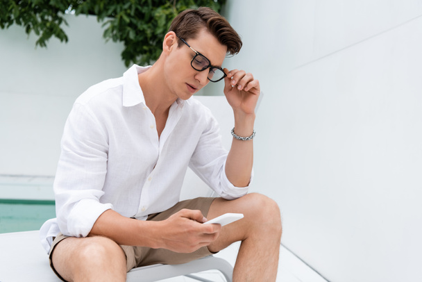 man adjusting eyeglasses while chatting on cellphone outdoors - Photo, Image