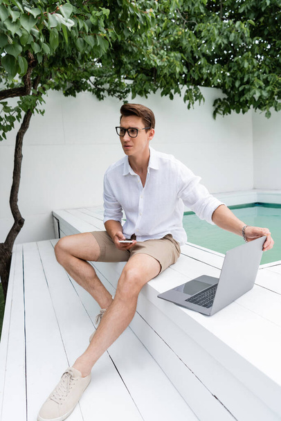 full length of man with laptop and mobile phone sitting near pool under green branches - Фото, изображение