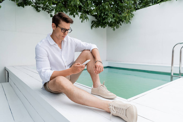 full length of man in summer clothes and eyeglasses messaging on smartphone near pool - Foto, Imagen