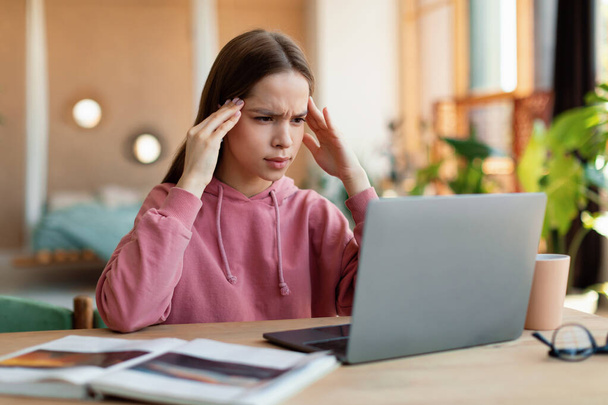 Uitgeput schoolmeisje met hoofdpijn, overwerken, te veel online huiswerk, achter het bureau zitten en naar een laptop kijken. Moe adolescent geconfronteerd met moeilijkheden met studeren op afstand - Foto, afbeelding