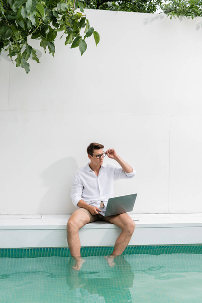 man in white shirt sitting near pool with laptop and looking away - Photo, Image