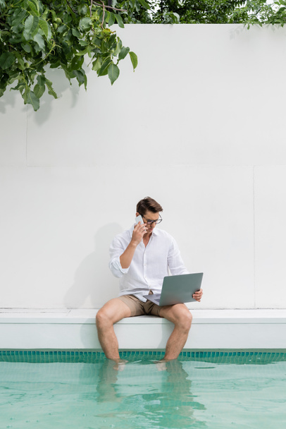 чоловік сидить з ногами в басейні і розмовляє на мобільному телефоні біля ноутбука
 - Фото, зображення