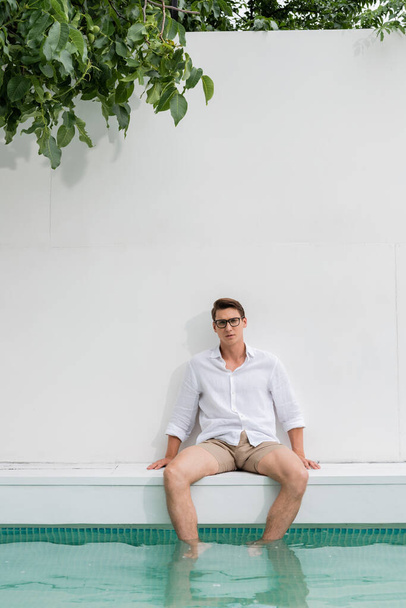 man in shirt and eyeglasses sitting near pool by white wall - Foto, Bild