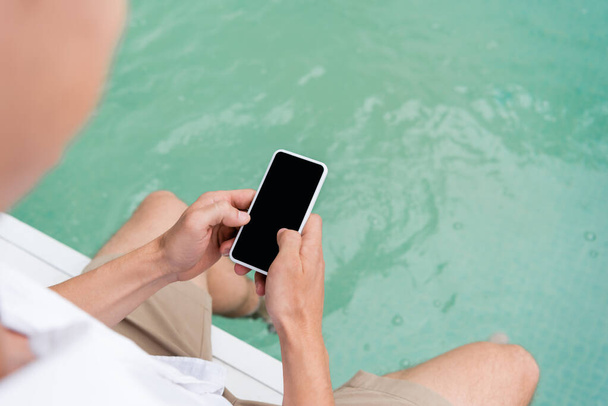 cropped view of man messaging on smartphone with blank screen near pool with turquoise water  - 写真・画像