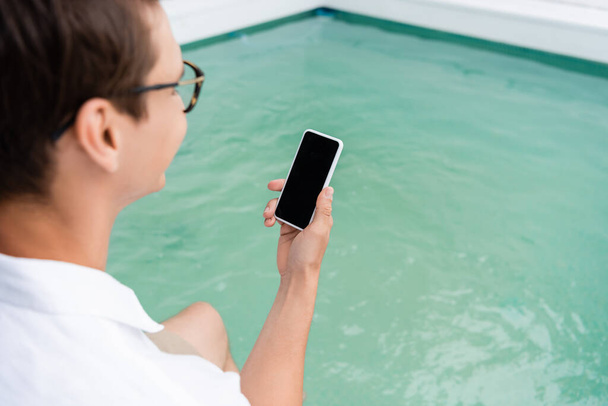 wazige man houdt mobiele telefoon met leeg scherm in de buurt van wazig zwembad - Foto, afbeelding