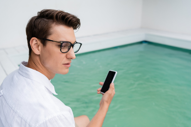 uomo in occhiali che tiene smartphone con schermo bianco vicino alla piscina sfocata - Foto, immagini