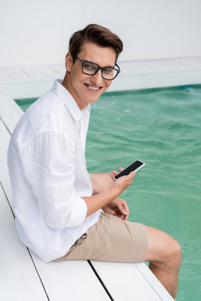 joyful man in eyeglasses sitting near pool and holding smartphone with blank screen - Photo, image