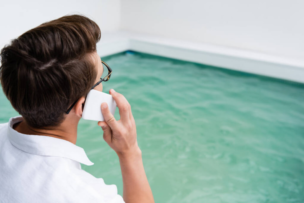 man met bril in gesprek op mobiele telefoon in de buurt van wazig zwembad met turquoise water - Foto, afbeelding