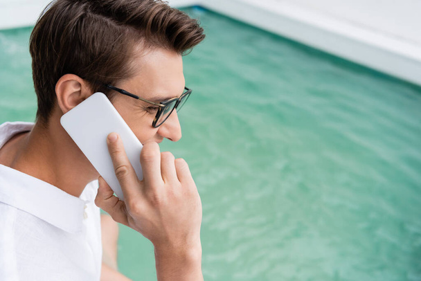 positive man in eyeglasses talking on mobile phone near pool with turquoise water - Photo, Image