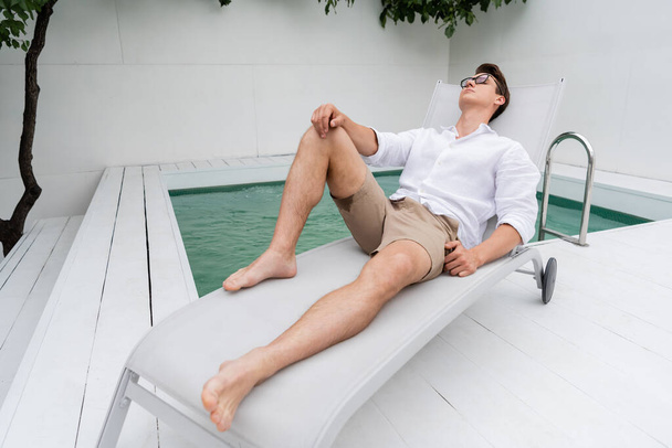 full length of barefoot man in summer clothes resting on deck chair at poolside - Photo, Image