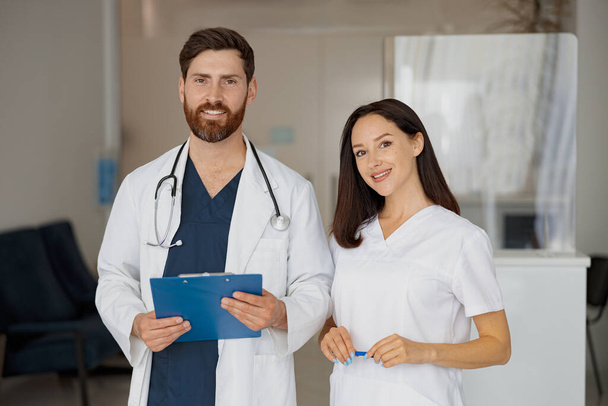 Retrato de médico y enfermera de pie cerca de la recepción en el hospital y mirando a la cámara. Foto de alta calidad - Foto, Imagen