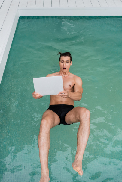top view of excited man with open mouth falling in pool with laptop - Fotoğraf, Görsel