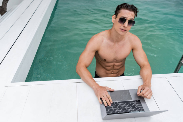 Sportlicher Mann mit Sonnenbrille mit Laptop am Pool - Foto, Bild