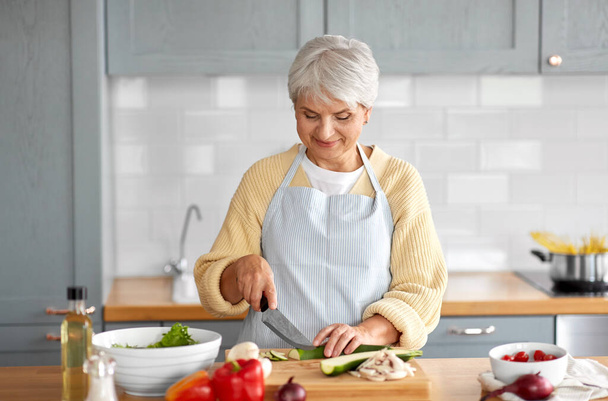 zdravé stravování, jídlo vaření a kulinářské pojetí - šťastná usměvavá seniorka s nožem sekání cukety na kuchyni doma - Fotografie, Obrázek