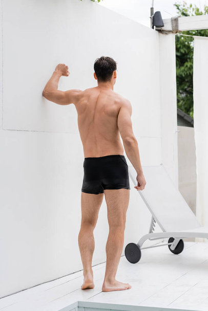 back view of wet muscular man in swimming trunks near white wall and deck chair - Фото, изображение