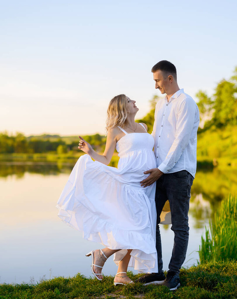 Beautiful young pregnant woman with her husband in the forest at sunset on the background of the lake - Foto, Bild