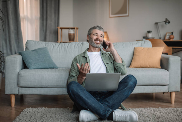 Усміхнений зрілий чоловік з бородою працює вдома, дзвонить клієнту по телефону з ноутбуком в інтер'єр вітальні. Сучасний гаджет для бізнесу, соціальної відстані та фрілансу для пенсіонерів
 - Фото, зображення